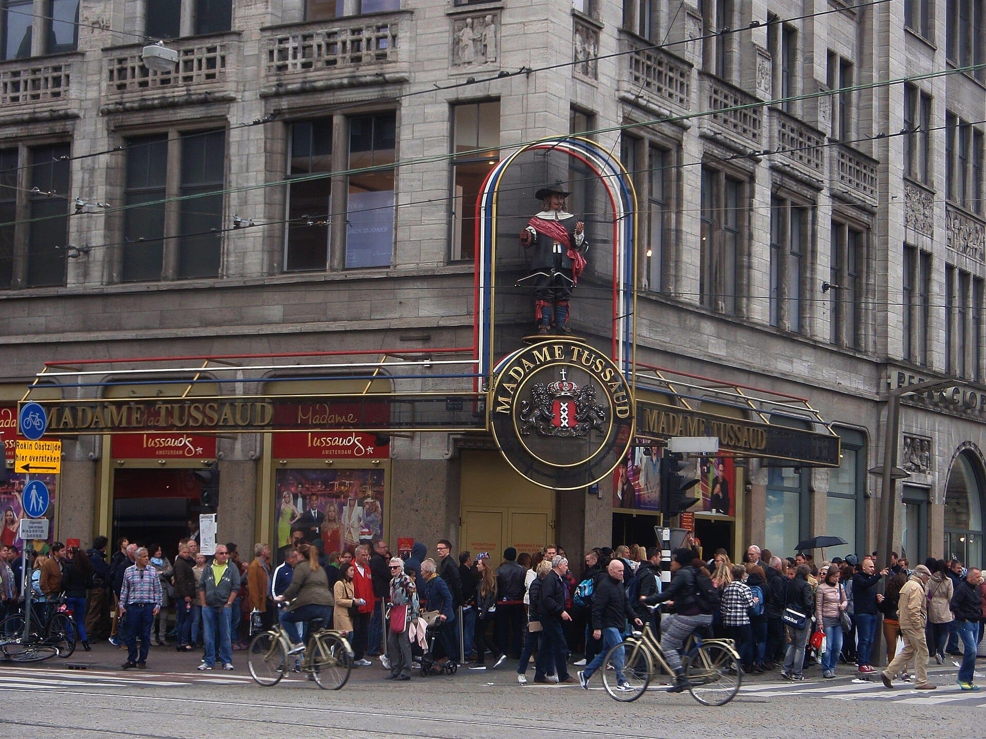 Menschenmengen warten vor dem Eingang von Madame Tussauds Amsterdam, einem historischen Gebäude mit großen Fenstern und einem markanten Wachsfigurenschild über der Tür.