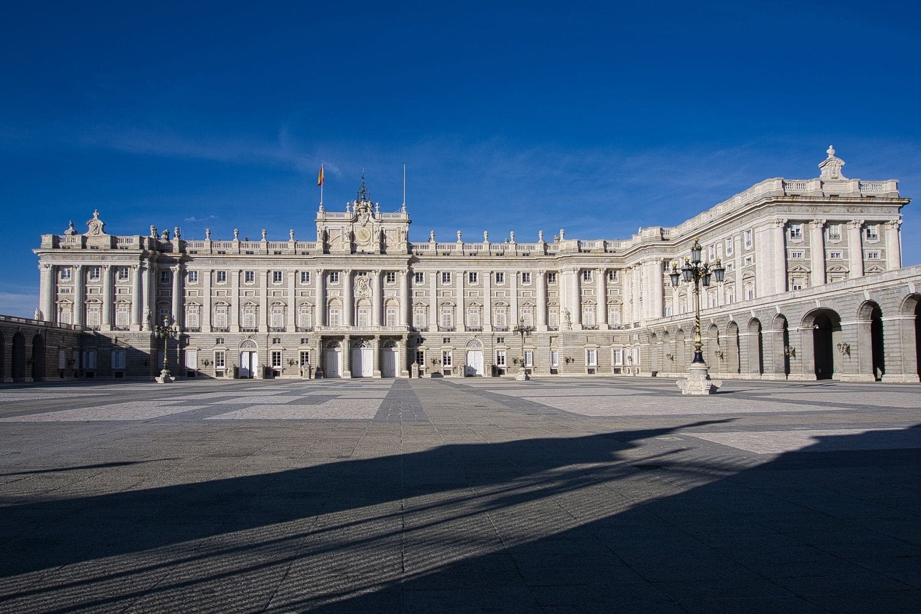 Wide view of Madrid's Royal Palace with its Neoclassical facade and broad courtyard under a clear blue sky.