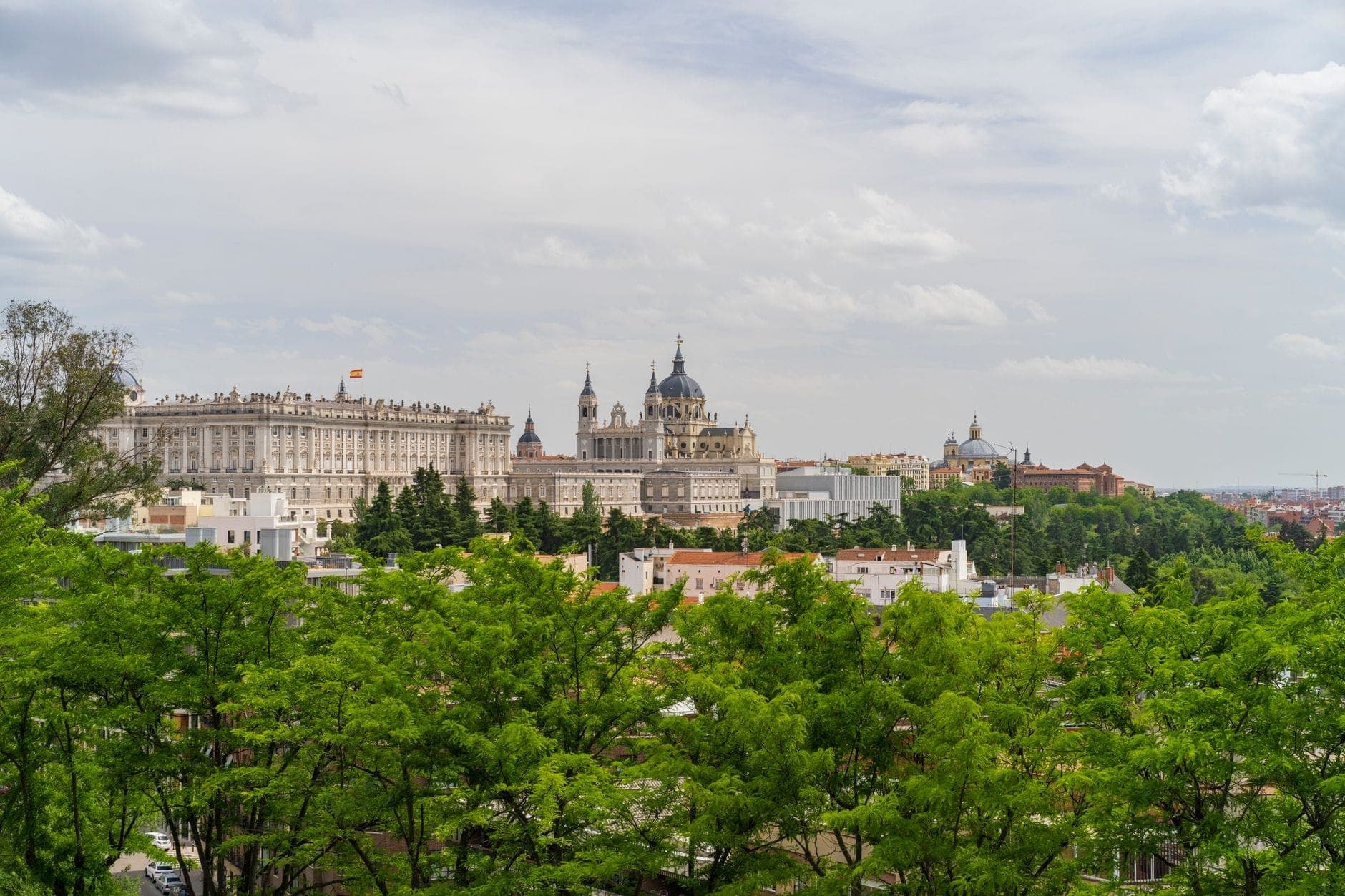 Panoramic view of Madrid showing the Almudena Cathedral and Royal Palace surrounded by gardens and greenery under a cloudy sky.