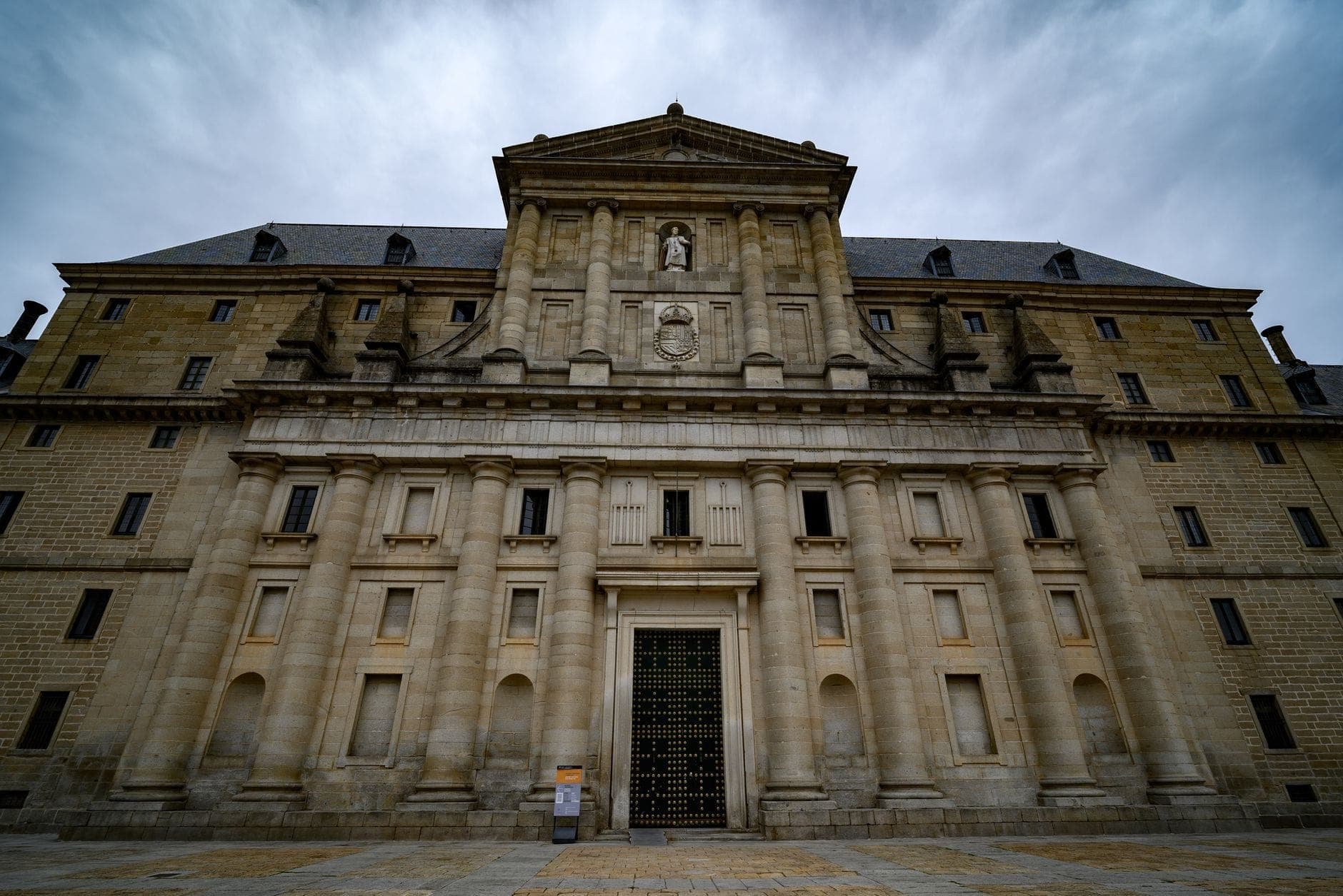 Stone facade of a grand historic monastery or convent in Madrid, with classical columns, niches, and a dramatic cloudy sky overhead.
