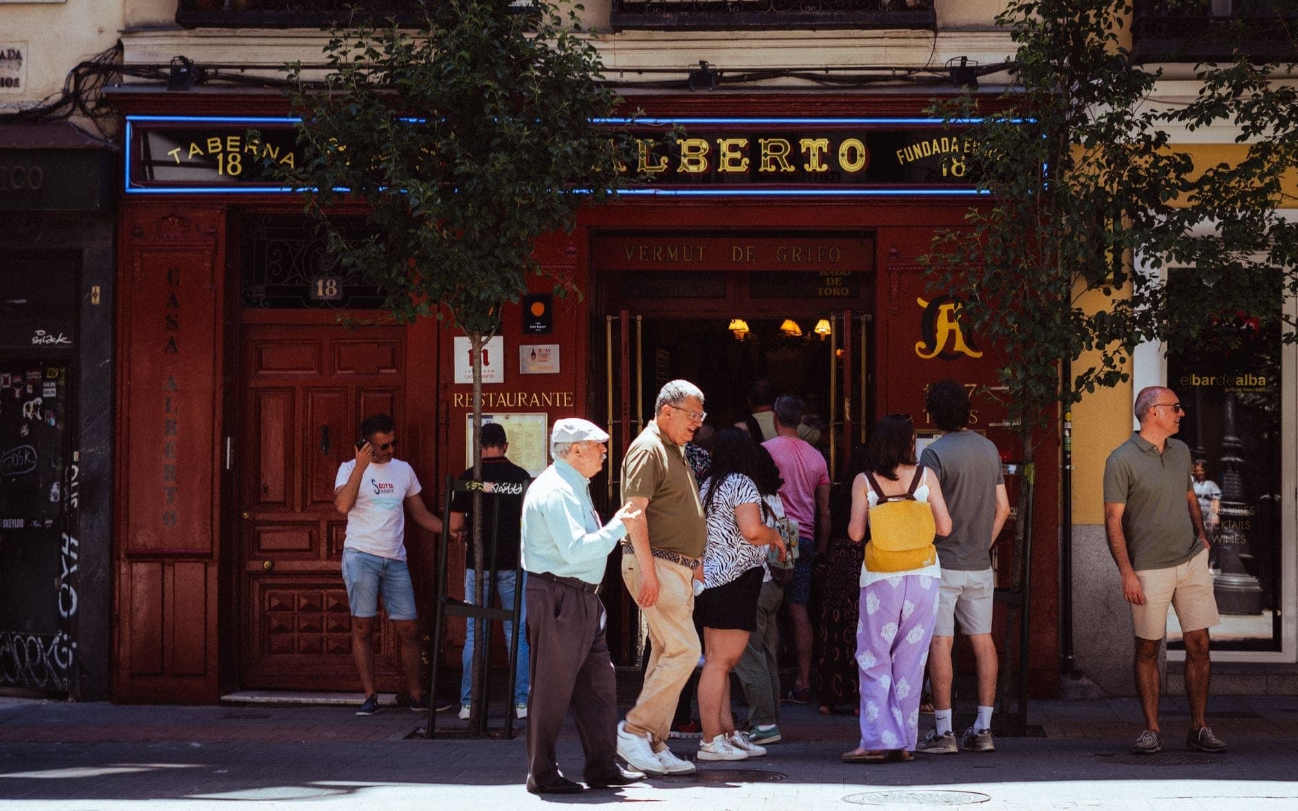 Ludzie zgromadzeni przed klasyczną restauracją w Madrycie w słoneczny dzień, z elewacją ozdobioną tradycyjnymi szyldami i ocienioną przez przydrożne drzewa.
