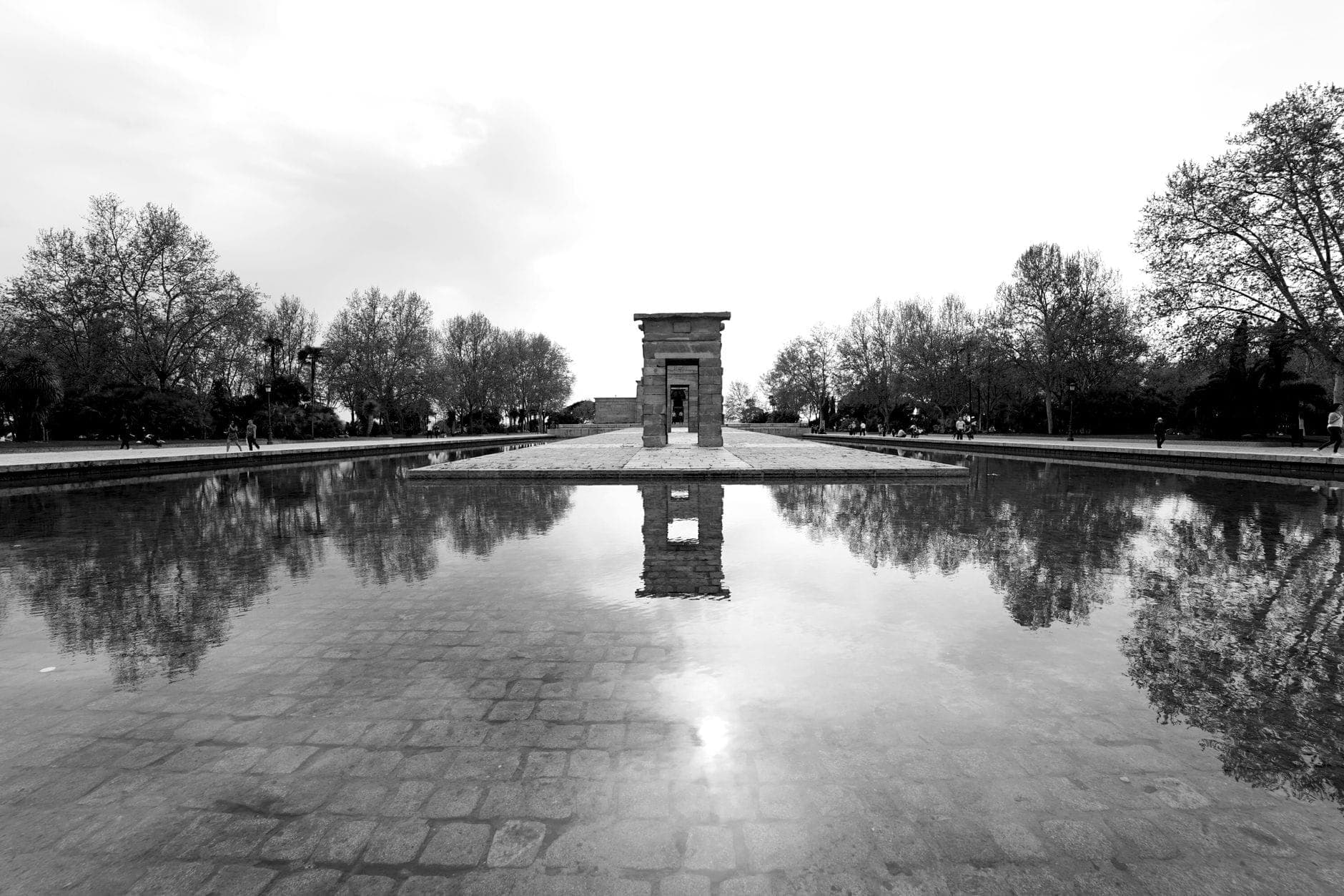 Foto en blanco y negro del Templo de Debod reflejado en un estanque, con árboles y cielo al atardecer al fondo.