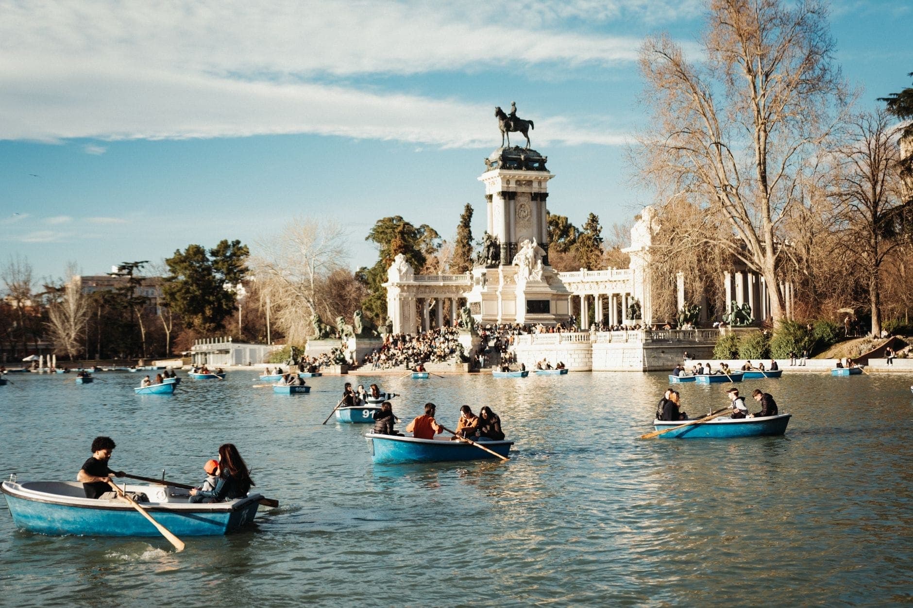 Parejas y grupos remando en botes azules en el gran lago del Parque del Retiro de Madrid, con el imponente monumento a Alfonso XII al fondo.