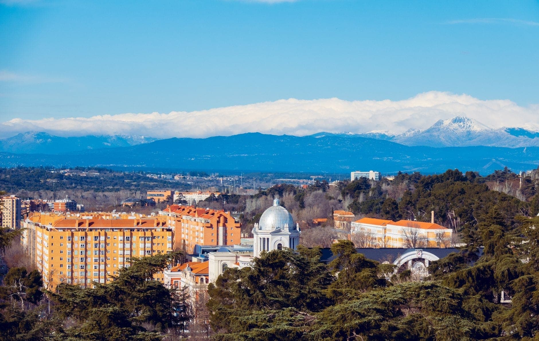 Wyraźny widok na budynki Madrytu z zabytkiem o białej kopule i ośnieżonymi górami Sierra de Guadarrama w tle pod błękitnym niebem.