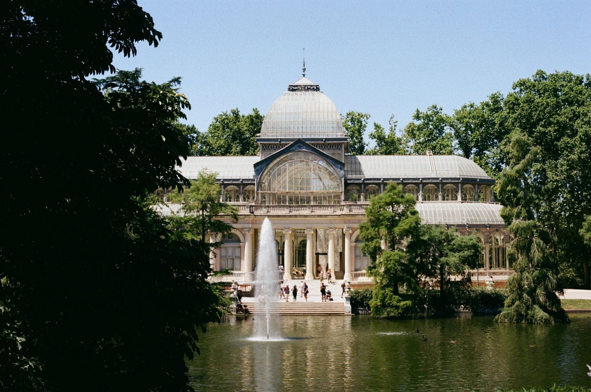 El Palacio de Cristal en el Parque del Retiro de Madrid con un lago, una fuente y frondosos árboles verdes bajo la luz de la primavera.