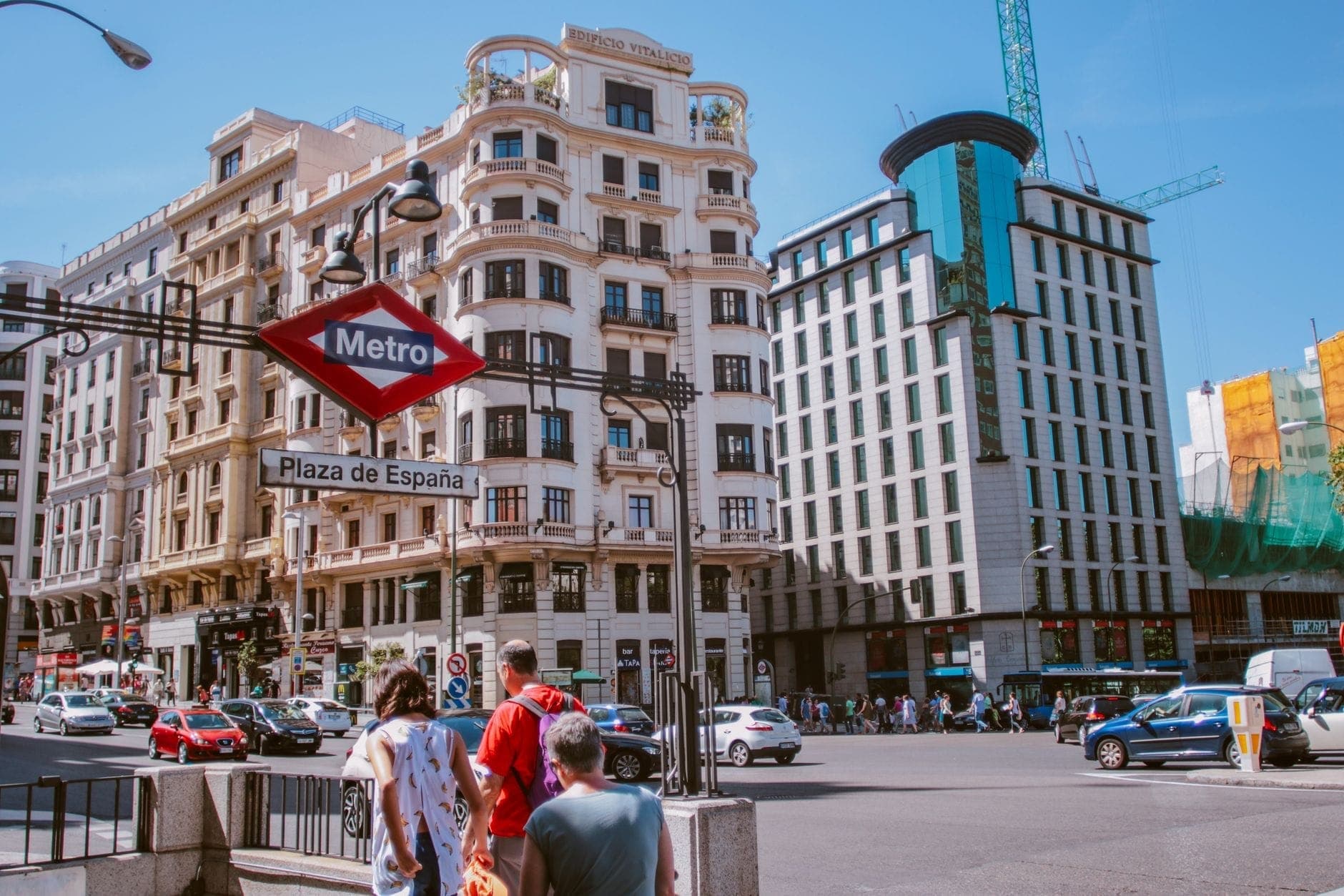 Personas bajando la entrada a la estación de Metro de Plaza de España en el centro de Madrid, con edificios de la ciudad y tráfico visibles en un día soleado.