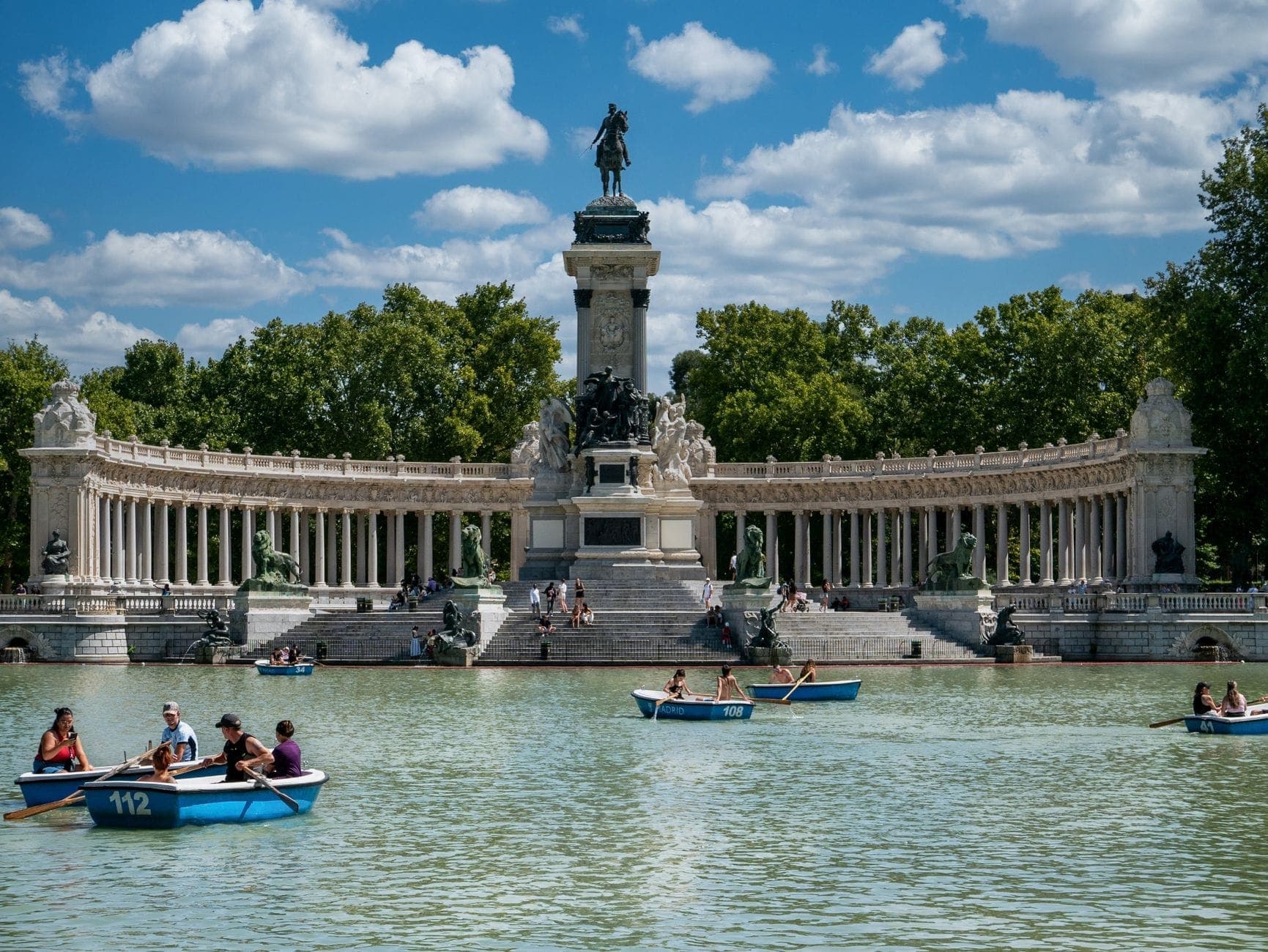 Monumento a Afonso XII no lago do Parque do Retiro em um dia ensolarado, pessoas remando em barcos azuis, árvores verdes ao fundo.