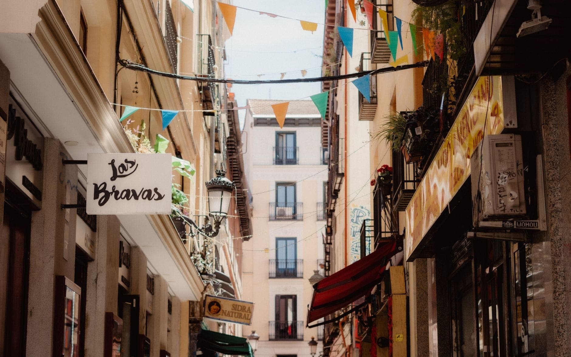 Uma rua estreita de Madrid decorada com bandeirolas festivas coloridas, letreiros externos e varandas, evocando a animada atmosfera das festas de bairro de verão.