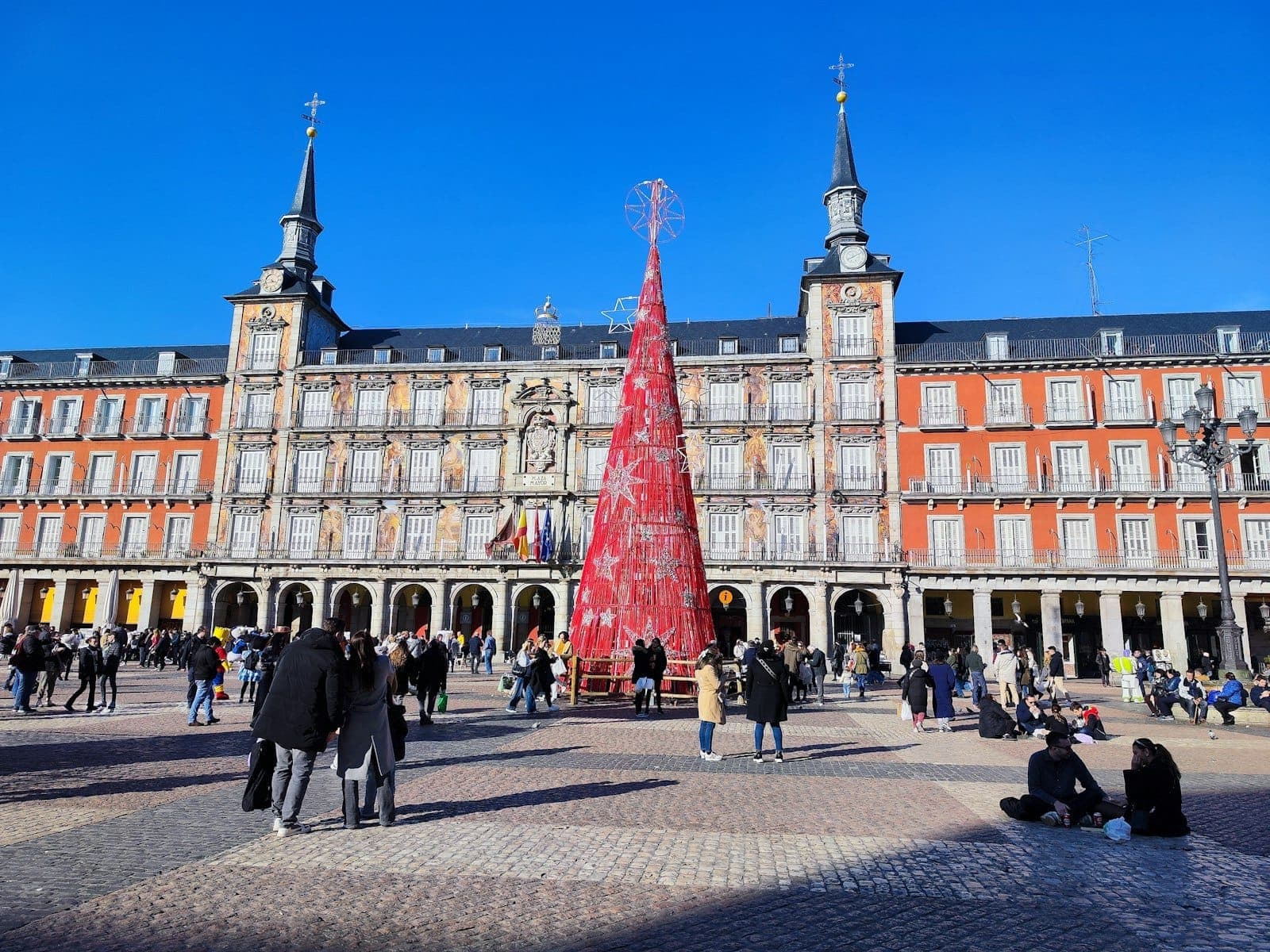 Persone riunite nella Plaza Mayor di Madrid decorata con un grande albero di Natale rosso al centro in una soleggiata giornata invernale.