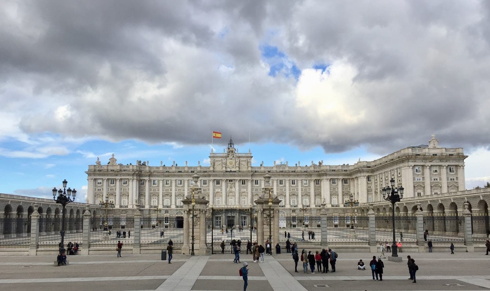 Vista ampia del Palazzo Reale di Madrid con persone nella piazza, sotto spettacolari nuvole invernali e una bandiera spagnola in cima all'edificio.