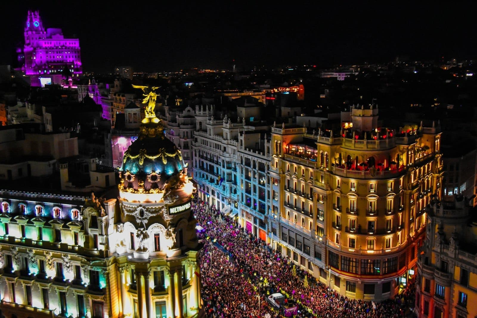 Gran Vía affollata a Madrid di notte con luci colorate, edifici storici e molte persone che festeggiano per strada.