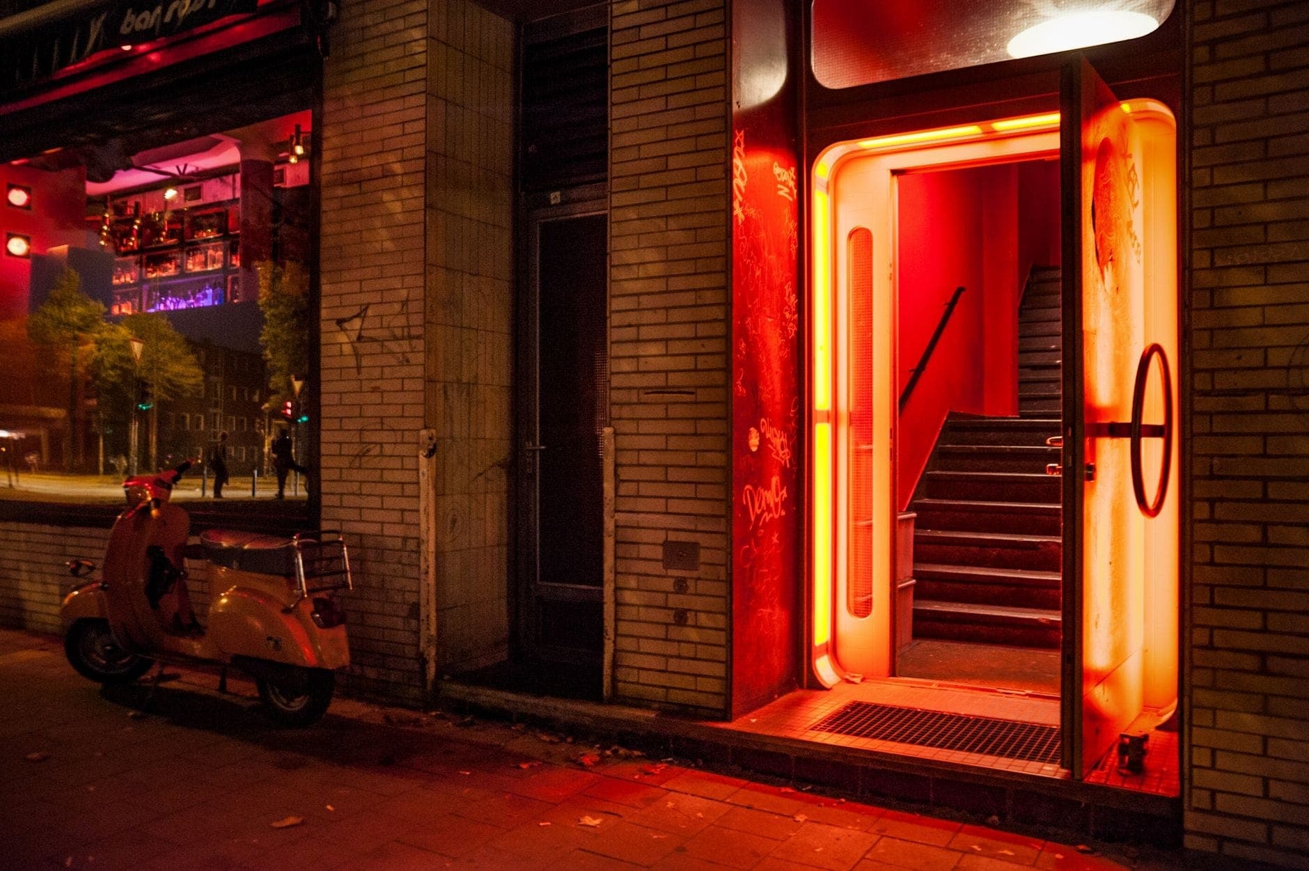 Ingresso di un bar o club calorosamente illuminato di notte con un bagliore al neon rosso e uno scooter parcheggiato fuori, scena di vita notturna invitante.
