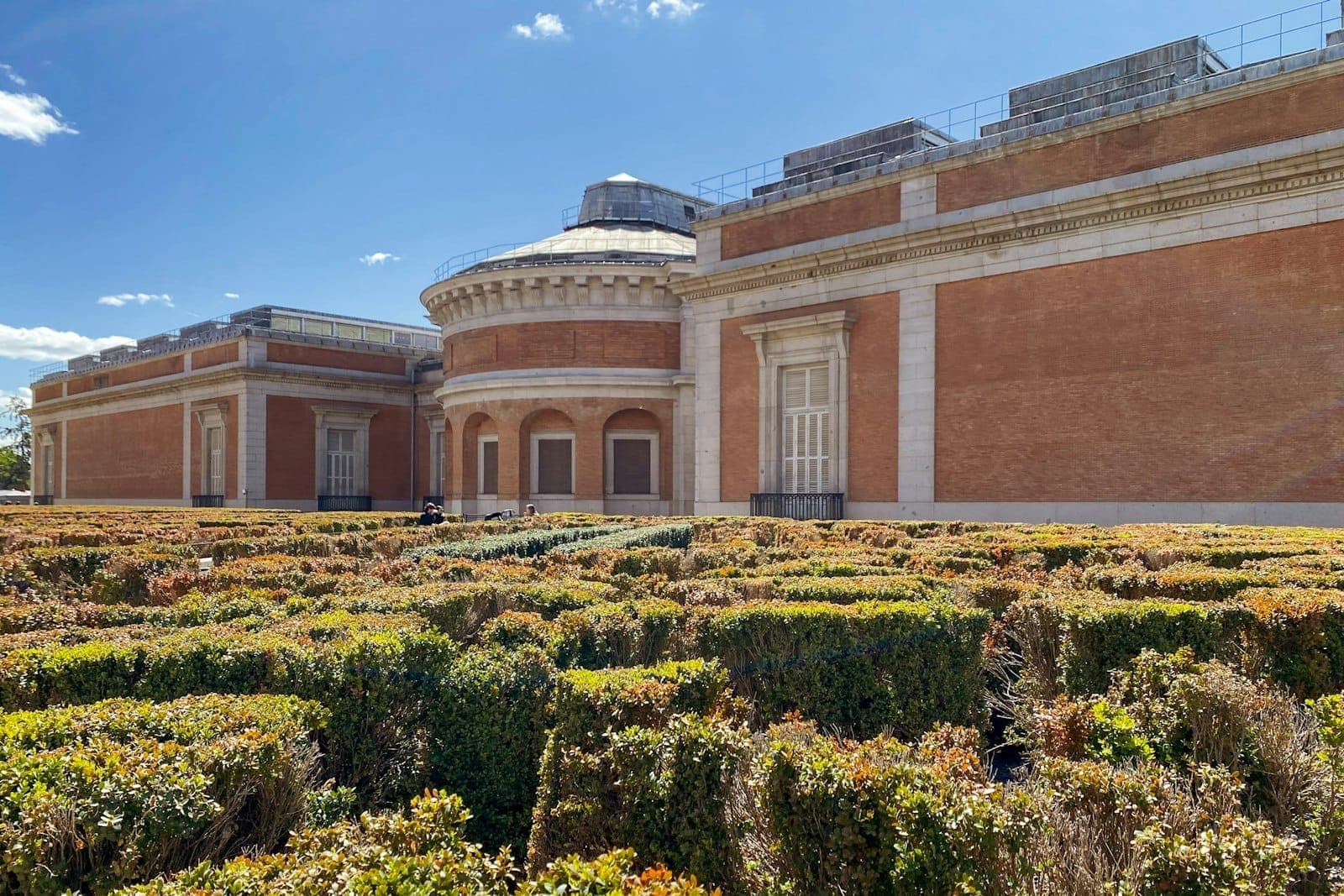 Vista esterna del Museo del Prado con la sua facciata in mattoni rossi e siepi ben rifilate in primo piano, sotto un cielo azzurro.