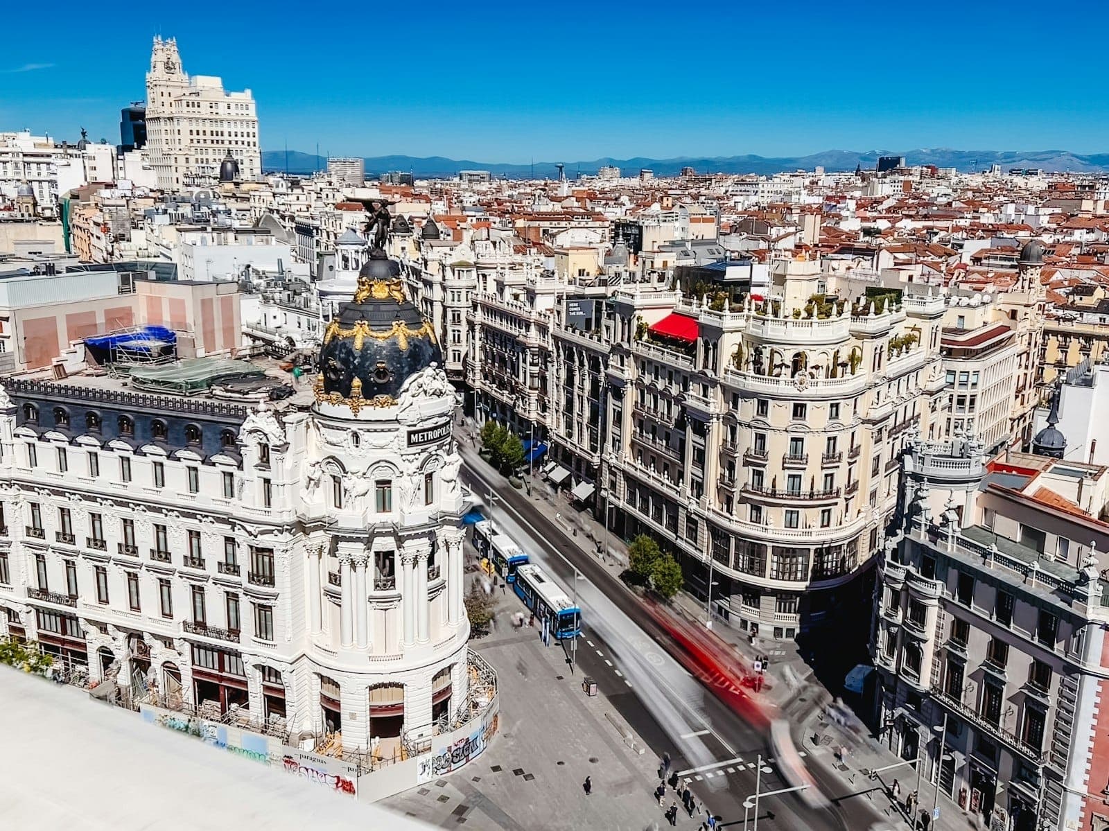 Panoramic view of Madrid’s iconic Gran Via with the Metropolis Building under a clear blue sky, busy streets, and cityscape in the background.