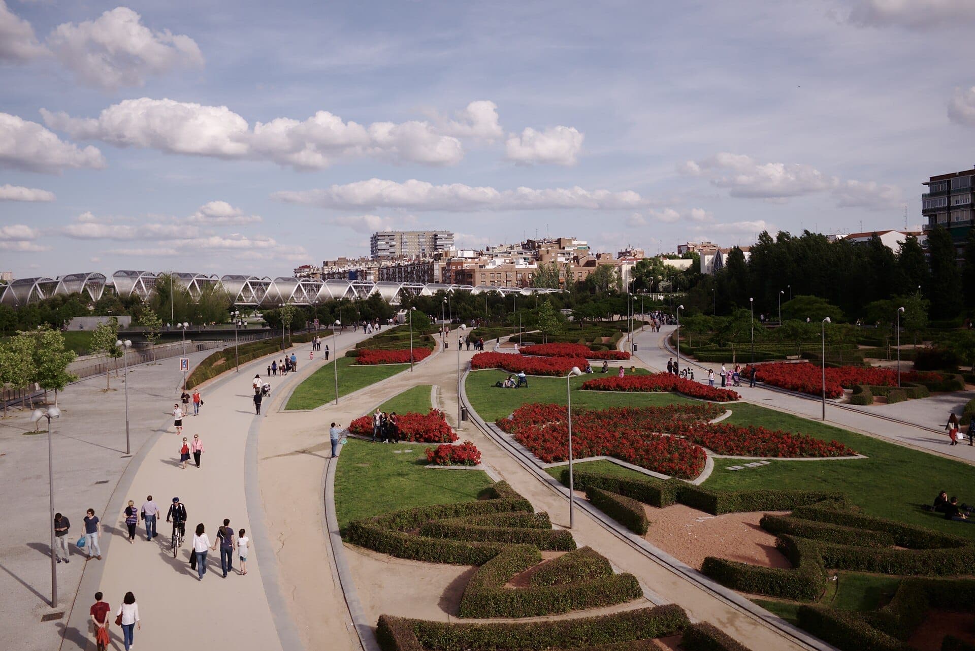 วิวกว้างของสวนสาธารณะ Madrid Río ที่มีสวนจัดภูมิทัศน์ สะพานคนเดินสมัยใหม่ ทางเดิน และผู้คนที่เพลิดเพลินกับบ่ายวันแดดจ้ากลางแจ้ง