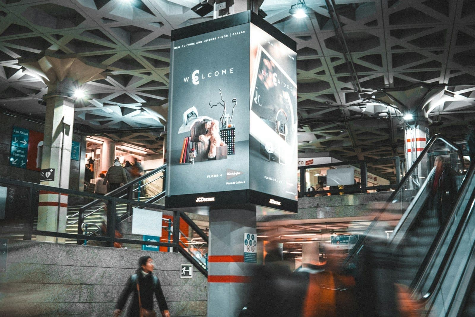 Belebtes Inneres einer U-Bahnstation in Madrid mit gehenden Menschen, Rolltreppen und großen Schildern über den Köpfen.