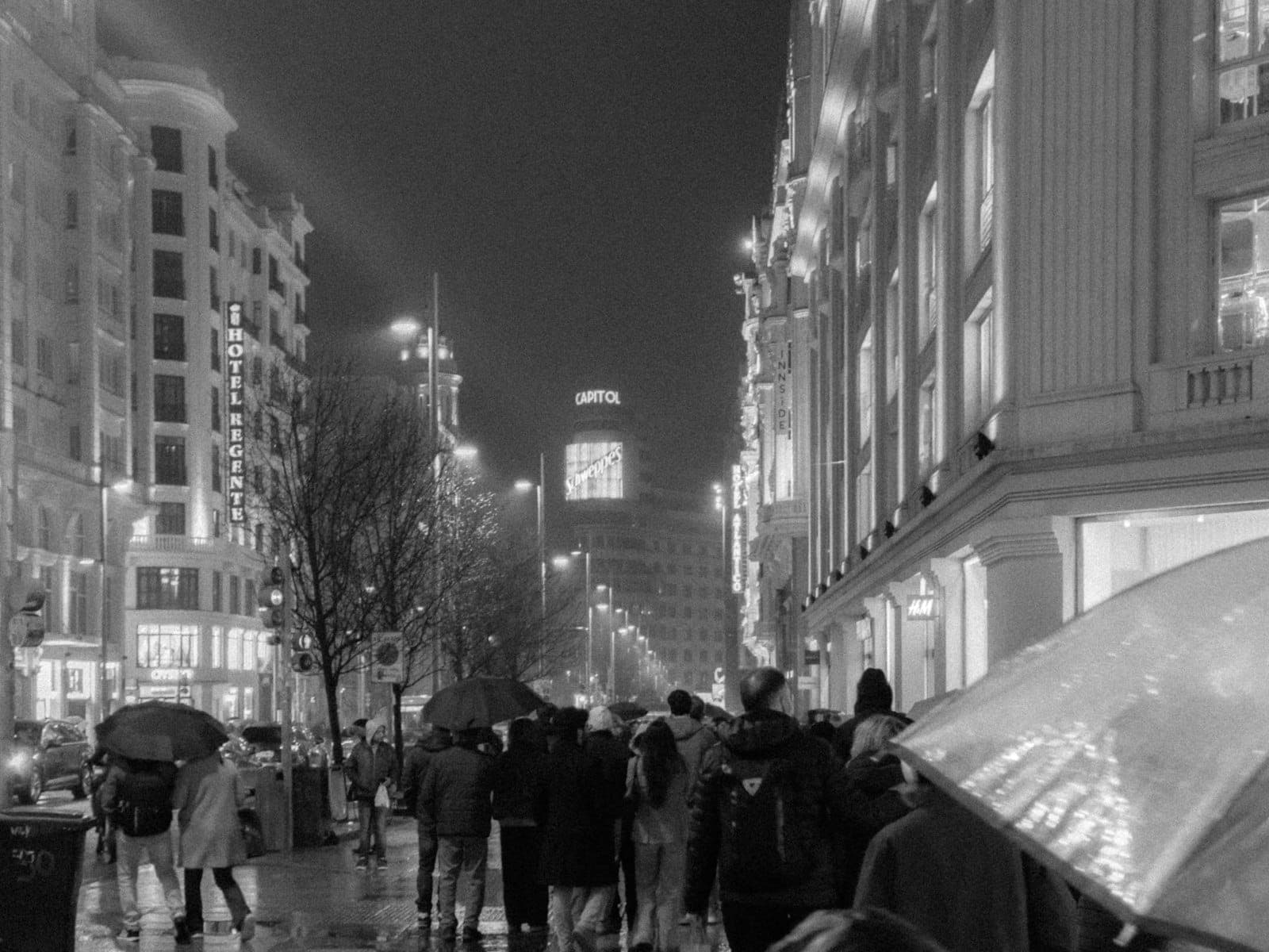 Belebte Straße in Madrid bei Nacht mit Menschen, die Regenschirme halten, glänzendem nassem Pflaster und ikonischen Gebäuden im Hintergrund.