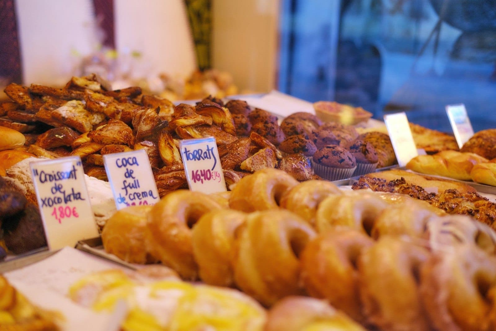 Primer plano de un escaparate de panadería con variados pasteles tradicionales etiquetados con nombres y precios en español, vistos a través del cristal de una tienda.