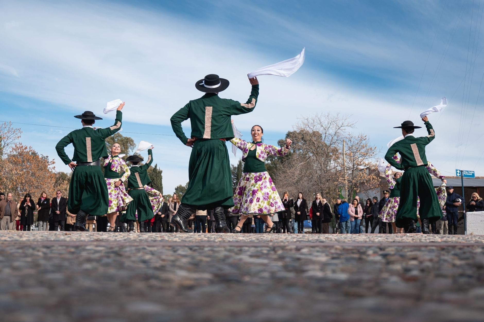 Grupo de bailarines con traje tradicional español actuando en una plaza al aire libre, observados por una multitud bajo un cielo azul.