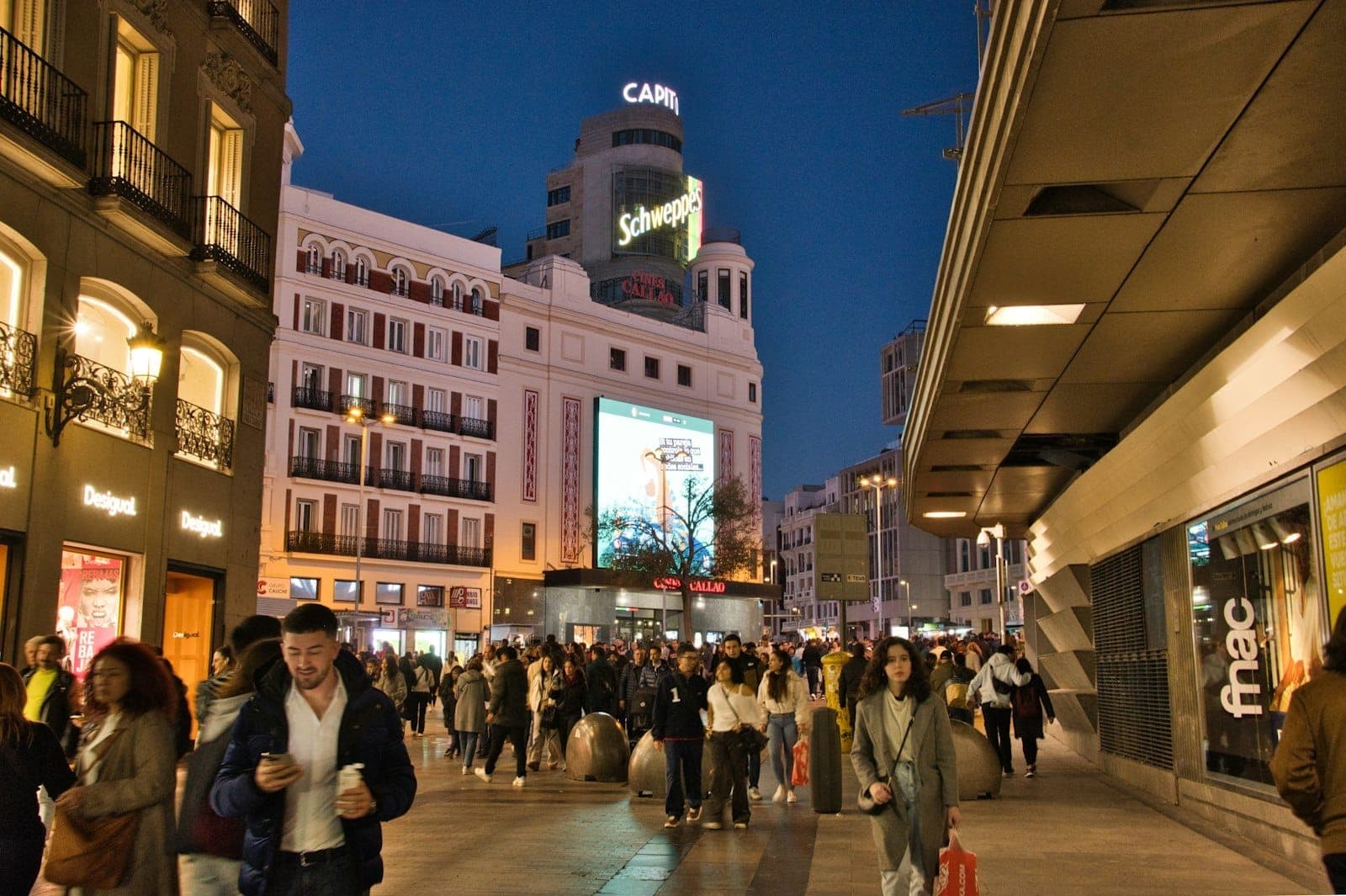 Belebte Einkaufsstraße in Madrid in der Abenddämmerung mit Einkaufsmassen, beleuchteten Schildern und markanten Gebäuden in der Nähe von Gran Vía und Sol.