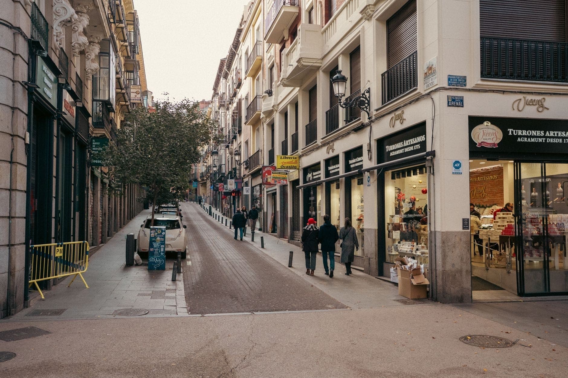 Schmale Einkaufsstraße in Madrid mit Fußgängern, gesäumt von modernen und älteren Gebäuden mit verschiedenen Geschäften und Cafés.