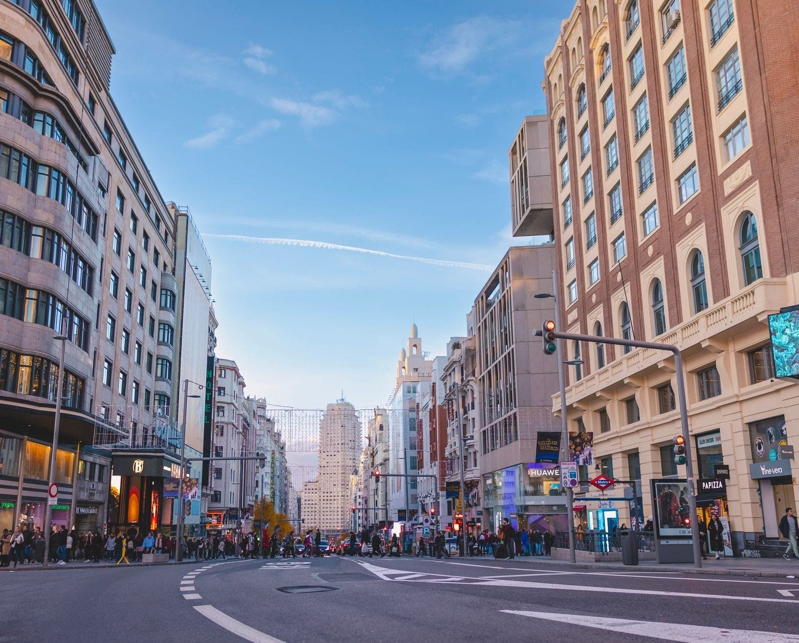 Weitwinkelansicht der Gran Vía in Madrid, mit Einkäufern, Verkehr und klassischen Gebäuden auf beiden Seiten dieser belebten Einkaufsstraße unter einem klaren blauen Himmel.