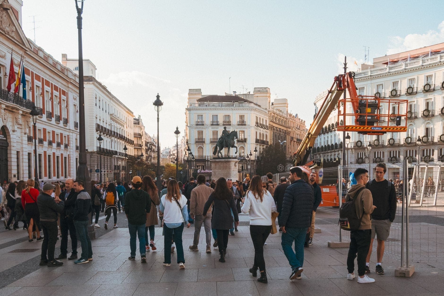Belebter Platz im Zentrum von Madrid mit Menschengruppen, historischen Gebäuden und einer Statue im Hintergrund bei Tageslicht.