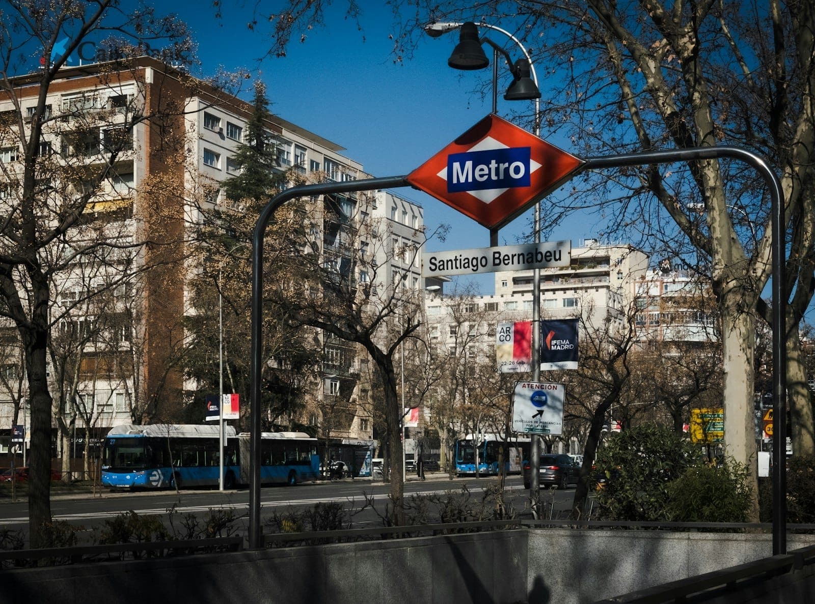 Eingang zur U-Bahnstation Santiago Bernabéu in Madrid mit Stadtgebäuden und Bussen im Hintergrund an einem klaren Tag