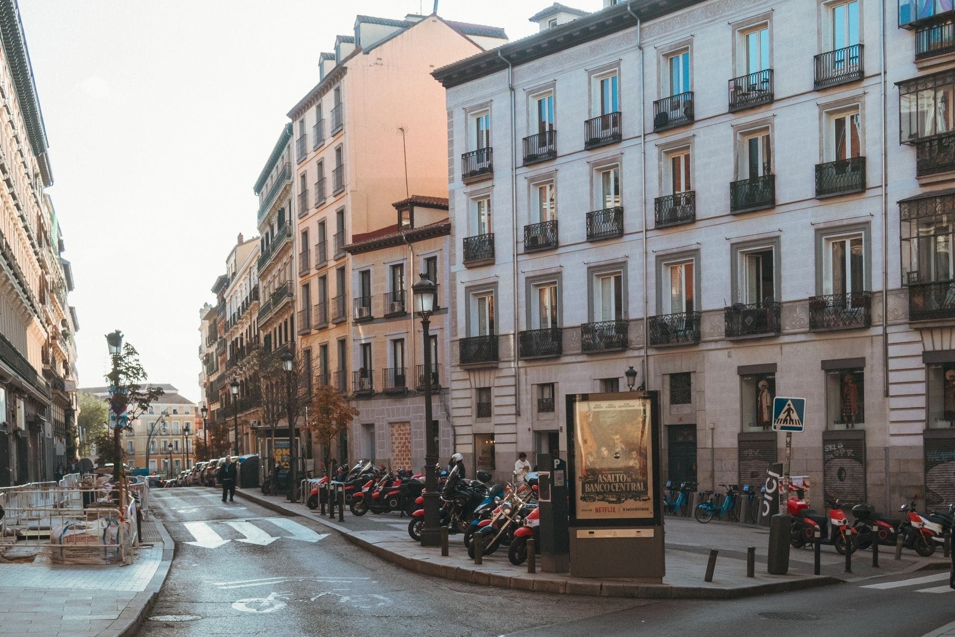 Weitwinkelansicht einer ruhigen Straße in Madrid, gesäumt von klassischen Wohngebäuden, am Gehweg geparkten Rollern und einigen Fußgängern.