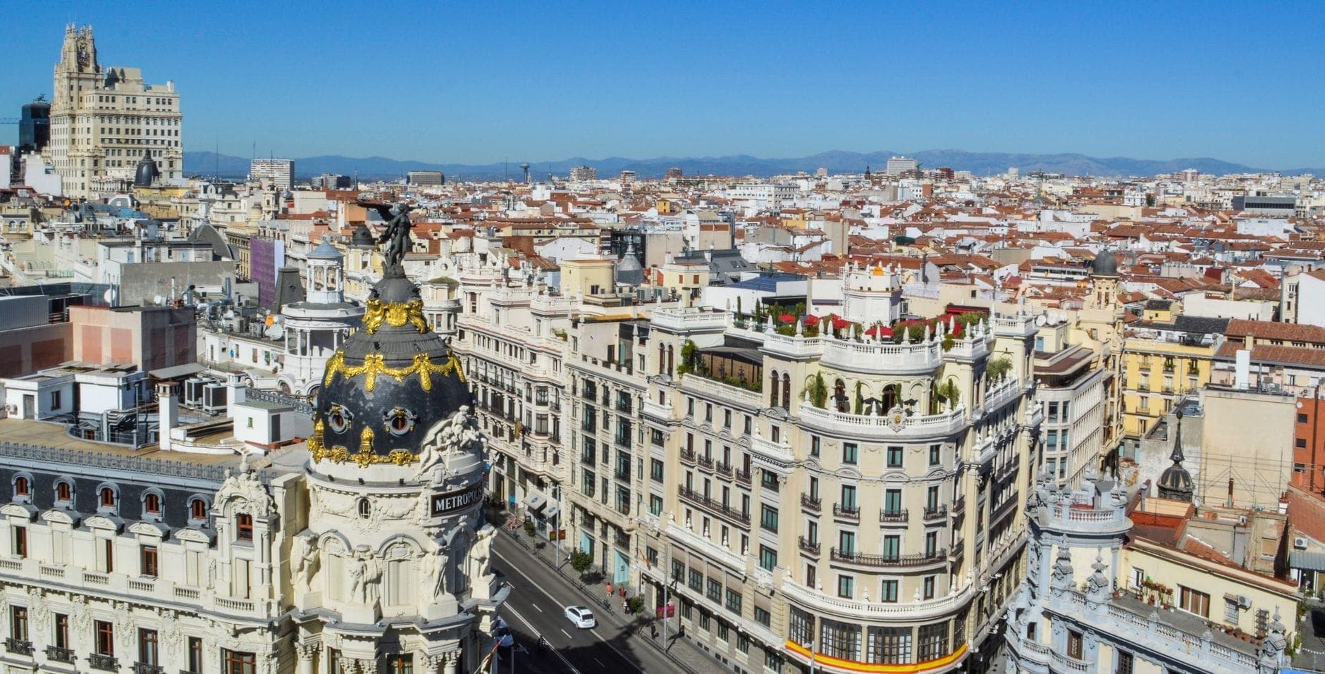 Helle Luftaufnahme der Gran Vía in Madrid und des Metrópolis-Gebäudes an einem sonnigen Tag mit klarem blauem Himmel und Bergen im Hintergrund.