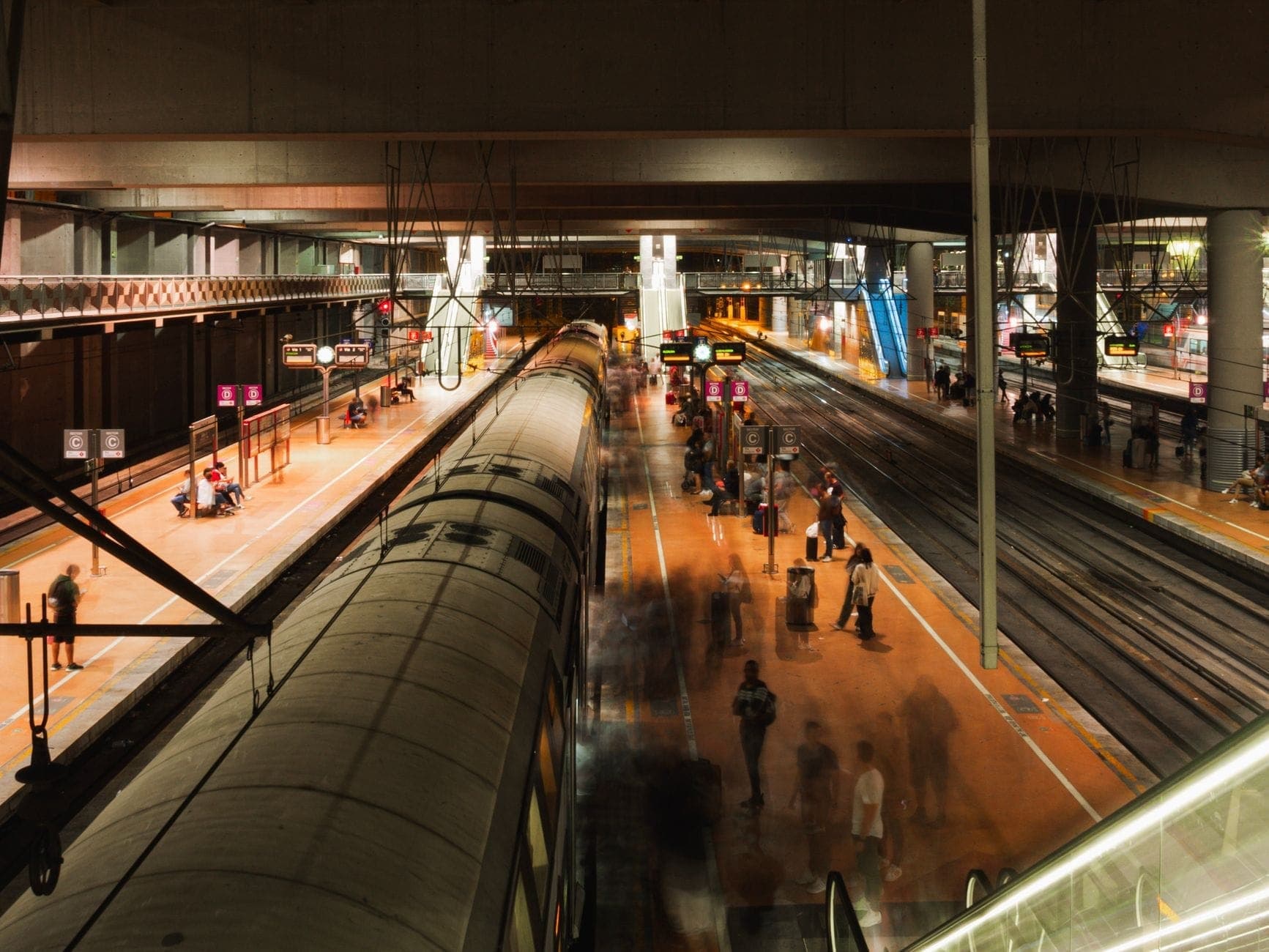 สถานีรถไฟที่คึกคัก มีผู้โดยสารเดินบนชานชาลาและรถไฟจอดบนหลายราง ภายใต้แสงไฟอบอุ่น