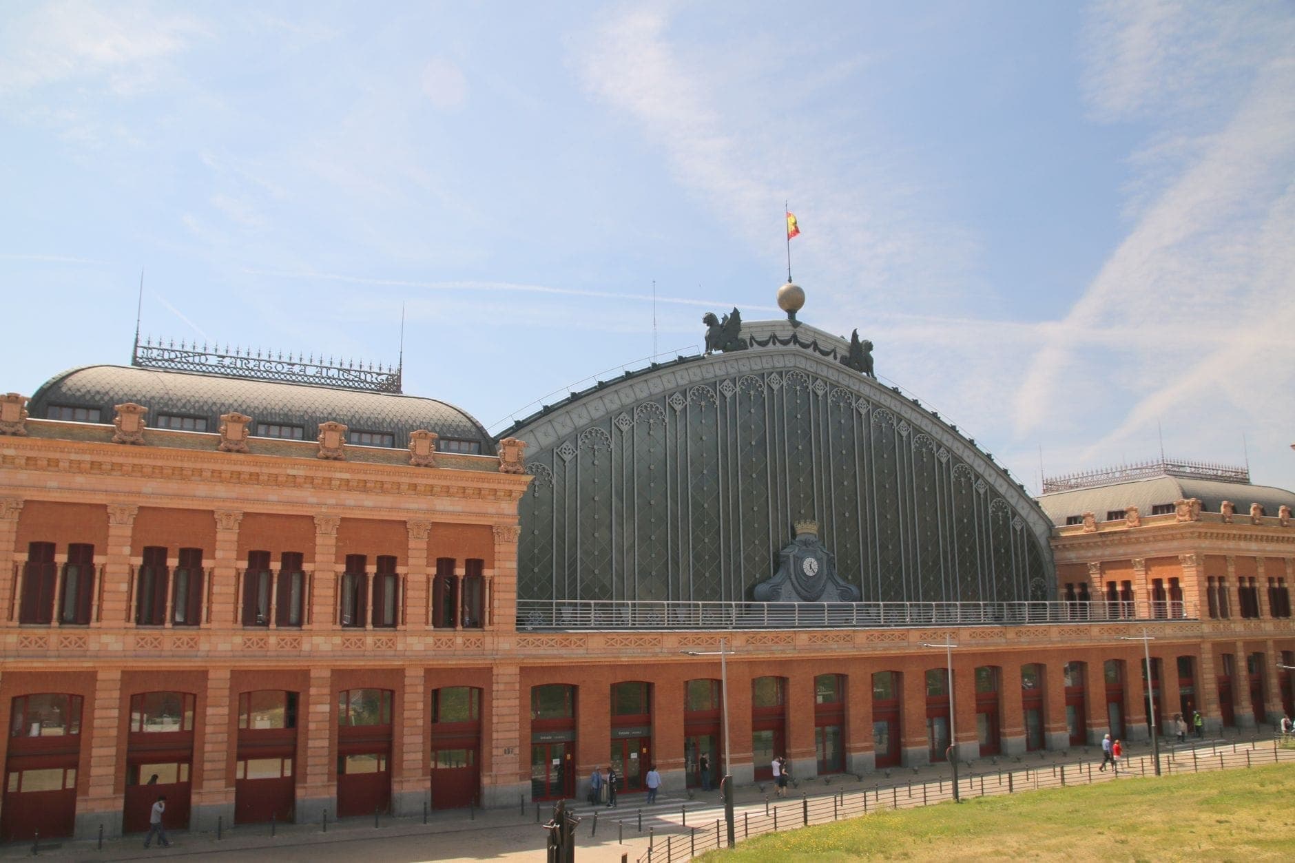 Die Fassade aus roten Ziegeln und Glas des Madrider Bahnhofs Atocha unter einem blauen Himmel, mit einer spanischen Flagge, die oben weht.