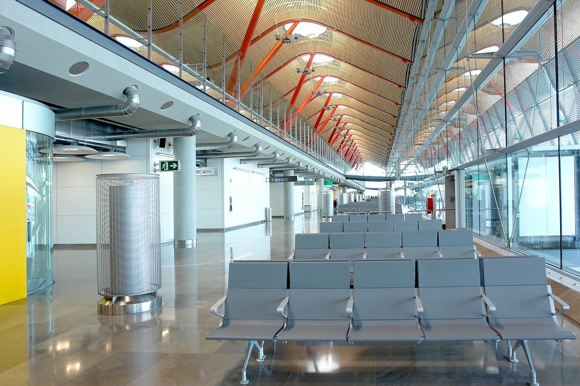 Sala de espera vacía con filas de asientos y el característico techo de madera y vigas rojas de la terminal del aeropuerto Madrid Barajas.