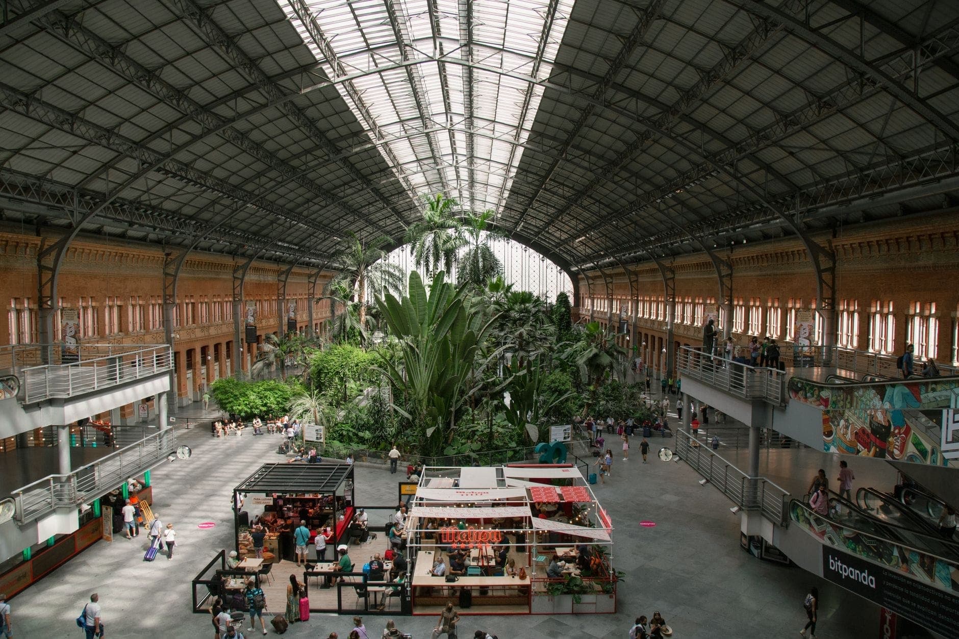 Interior de la estación de tren Atocha de Madrid con su distintivo techo de cristal, jardín tropical y personas caminando o sentadas en el amplio vestíbulo.