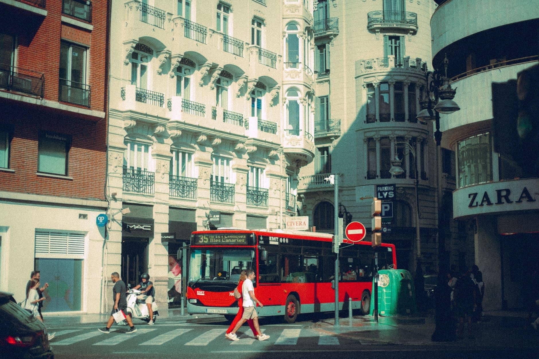 Un autobús rojo de la ciudad pasa por una intersección en el centro de Madrid con edificios históricos y peatones cercanos.