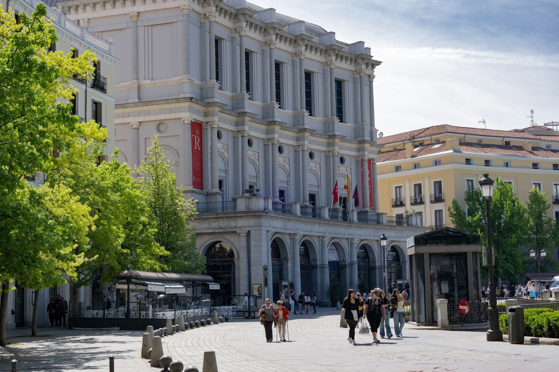 ผู้คนเดินเล่นใกล้โรงละครโอเปร่า Teatro Real ท่ามกลางวันแดดสดใส ต้นไม้เขียวและตึกแบบยุโรปคลาสสิกในฉากหลัง