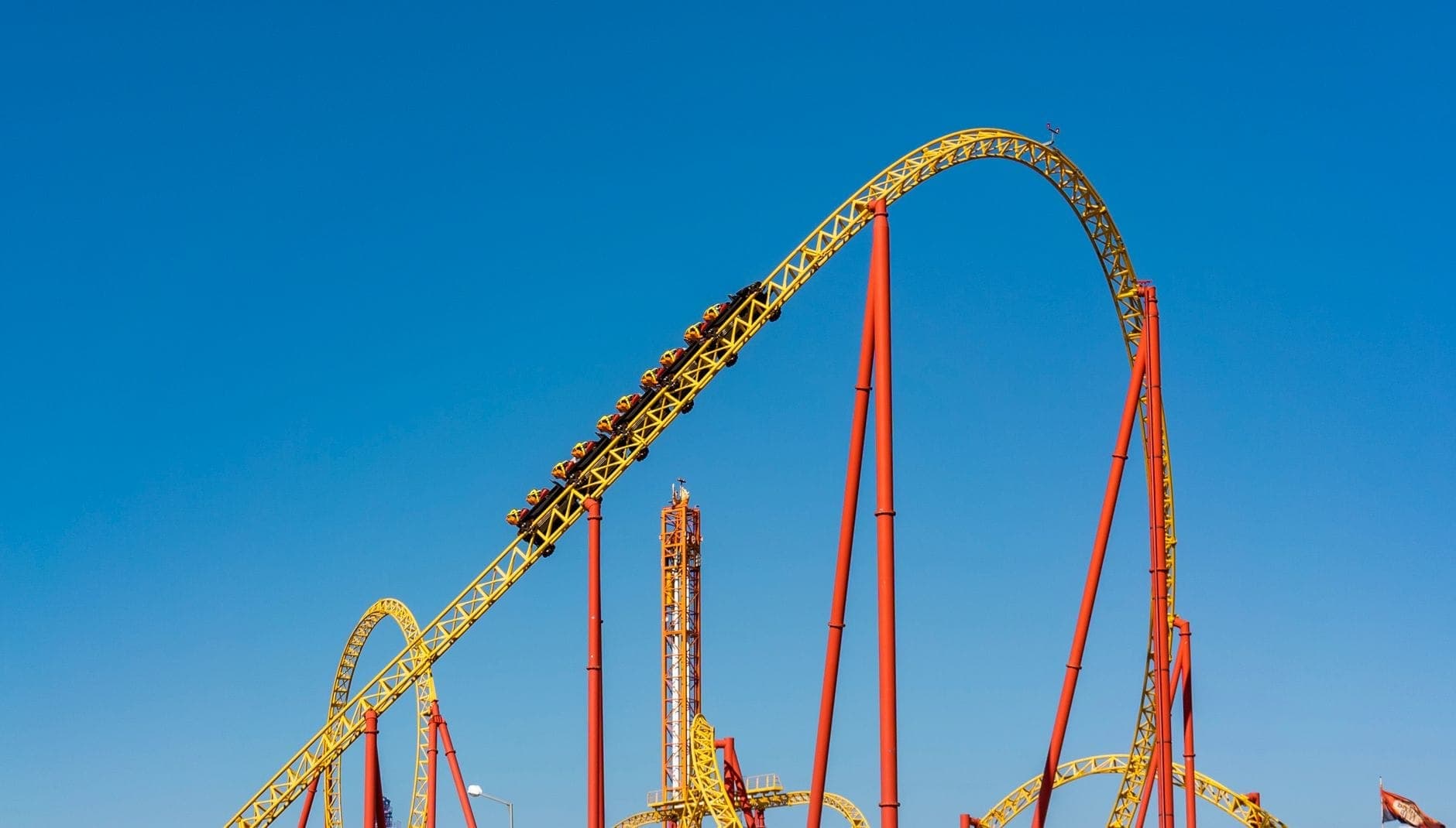Czerwono-żółty roller coaster na tle błękitnego nieba w parku rozrywki w Madrycie, wagony pełne pasażerów na szczycie stromego toru.