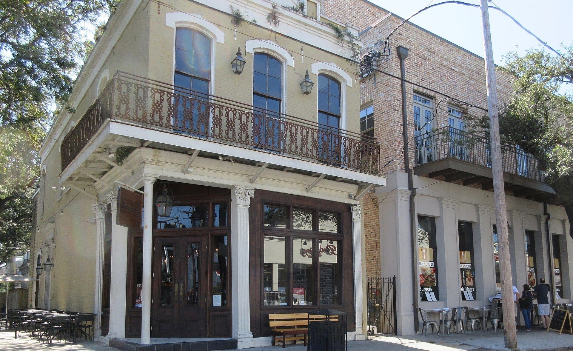 Historic brick buildings with balconies and sidewalk seating on Magazine Street in New Orleans, shaded by leafy trees on a sunny day.