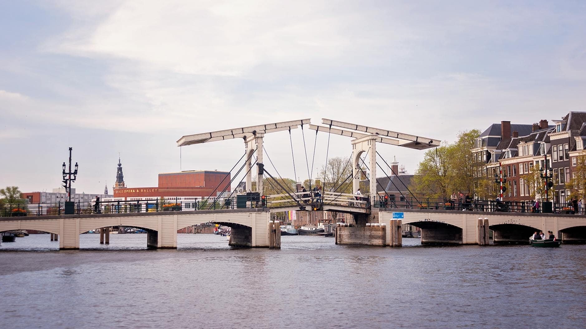 Weitwinkelansicht der weißen Klappbrücke Magere Brug über den Amstel in Amsterdam, umgeben von Gebäuden und Bäumen unter einem teils bewölkten Himmel.