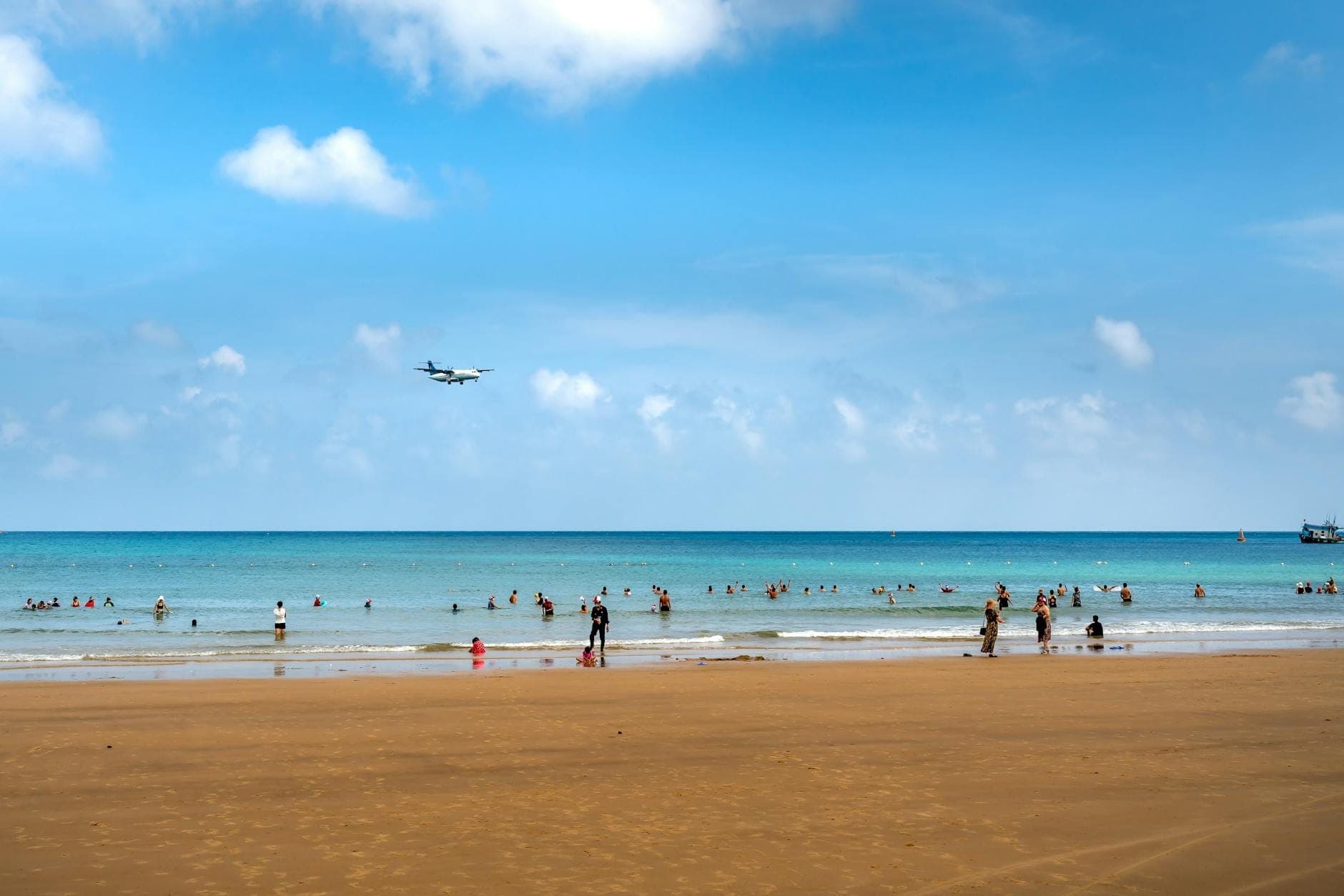 Wide view of Mai Khao Beach with soft sand, turquoise sea, scattered beachgoers, and an airplane flying low over the water under a bright blue sky.