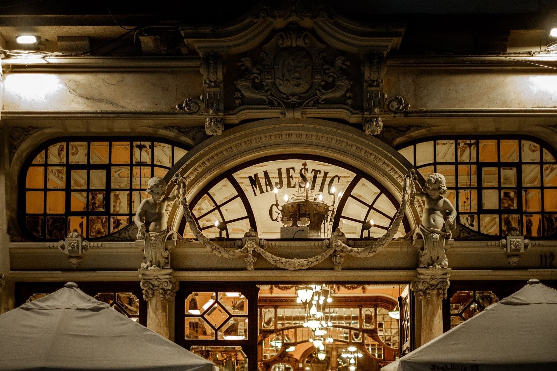 Zdobna fasada Café Majestic w Porto z rzeźbionymi cherubinami, wielkimi łukami i ciepło oświetlonymi lustrami odbijającymi wnętrze kawiarni w stylu Belle Époque.