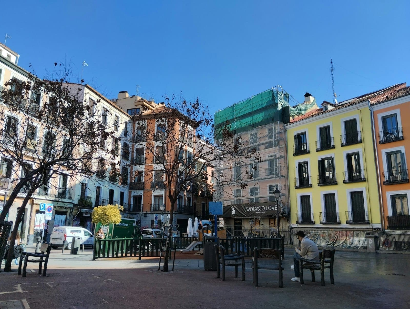 ลานกว้างกลาง Malasaña รายล้อมด้วยตึกที่พักสีสันสดใส ต้นไม้ไร้ใบ ม้านั่ง และท้องฟ้าสีฟ้าสดบนวันที่แดดจัด