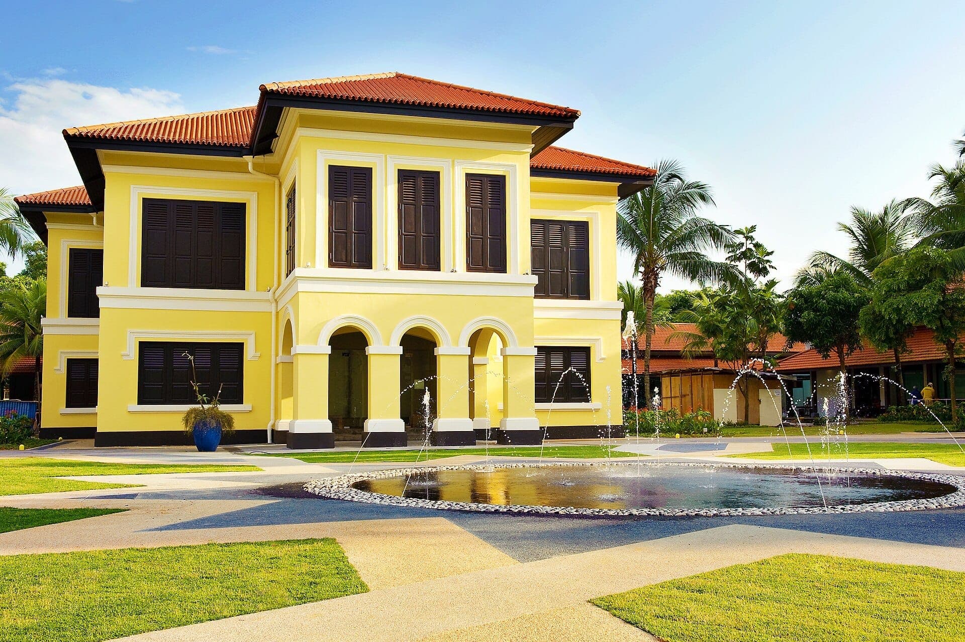 Malay Heritage Centre ในสิงคโปร์ อาคารอาณานิคมสีเหลืองสดใสพร้อมบานเกล็ดสีเข้ม ตั้งอยู่เบื้องหลังน้ำพุทรงกลมและสวนที่ตัดแต่งอย่างเรียบร้อยภายใต้ท้องฟ้าแจ่มใส