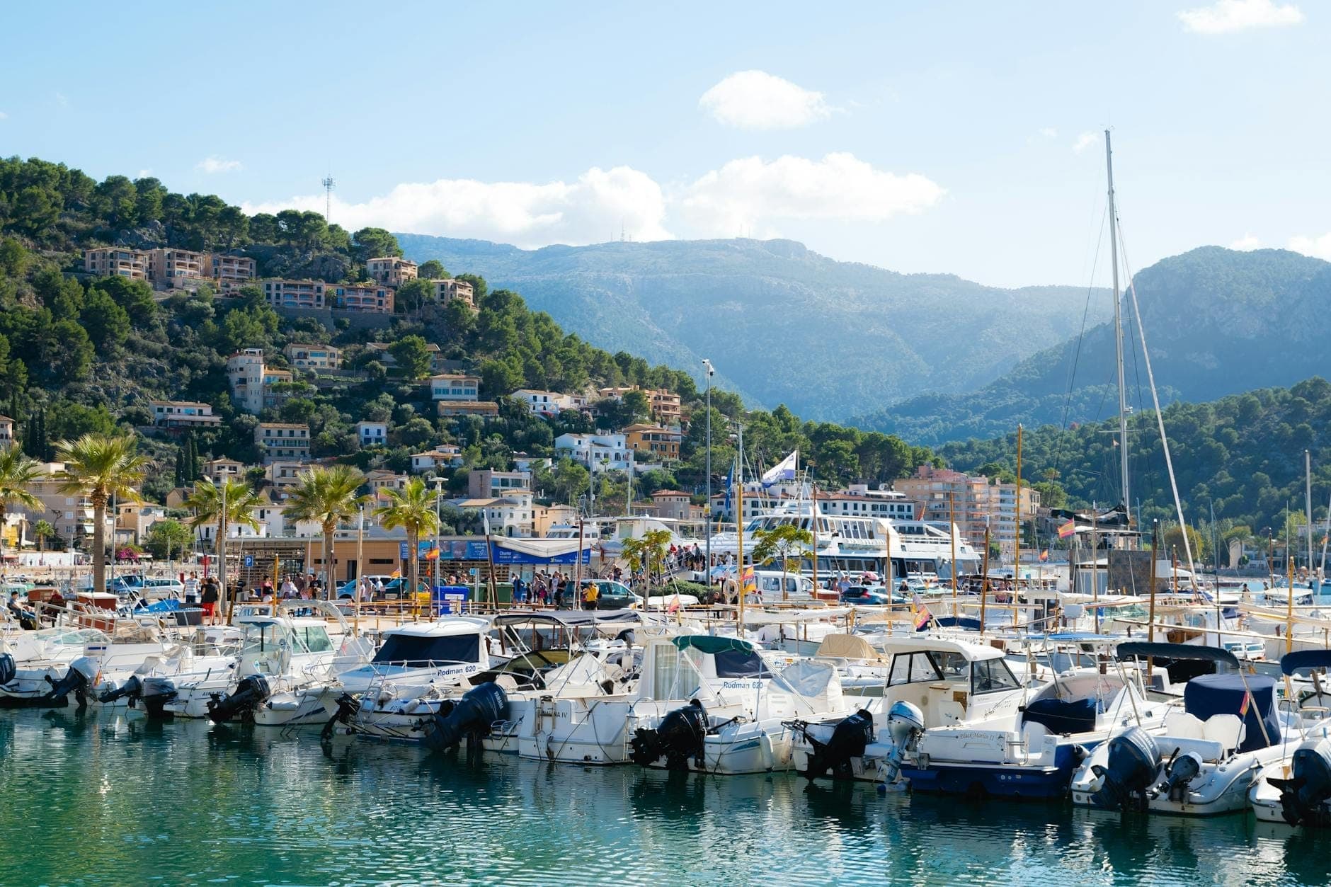 Marina auf Mallorca mit zahlreichen angedockten Booten, Gebäuden am Hang, Bergen im Hintergrund und klarem blauem Himmel.