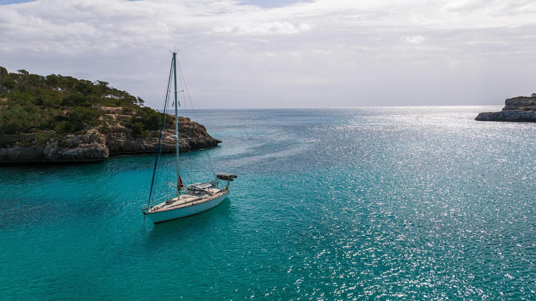 Ein einzelnes Segelboot liegt in einer atemberaubenden türkisfarbenen Bucht, eingerahmt von felsiger Küste und Pinien unter strahlend blauem Himmel – ein Bild für das entspannte Segelerlebnis auf Mallorca.