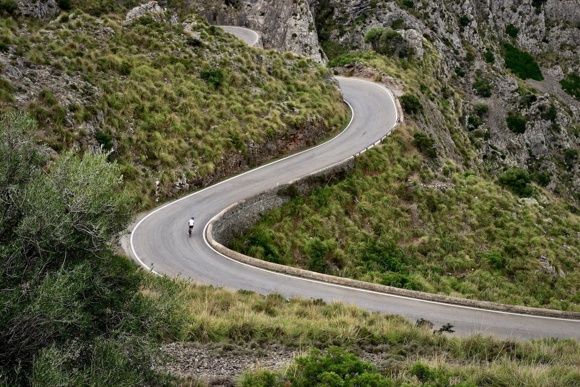 Ein einzelner Radfahrer erklimmt eine dramatische Spitzkehre auf einer kurvenreichen Bergstraße, umgeben von Felswänden und grünem Gras in der Serra de Tramuntana.