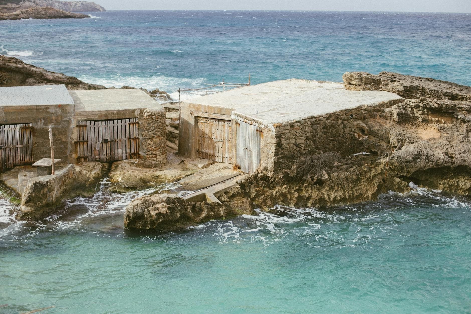 Cabañas de pesca tradicionales de madera en la orilla rocosa junto al mar turquesa cristalino en Mallorca, con olas salpicando en las entradas.