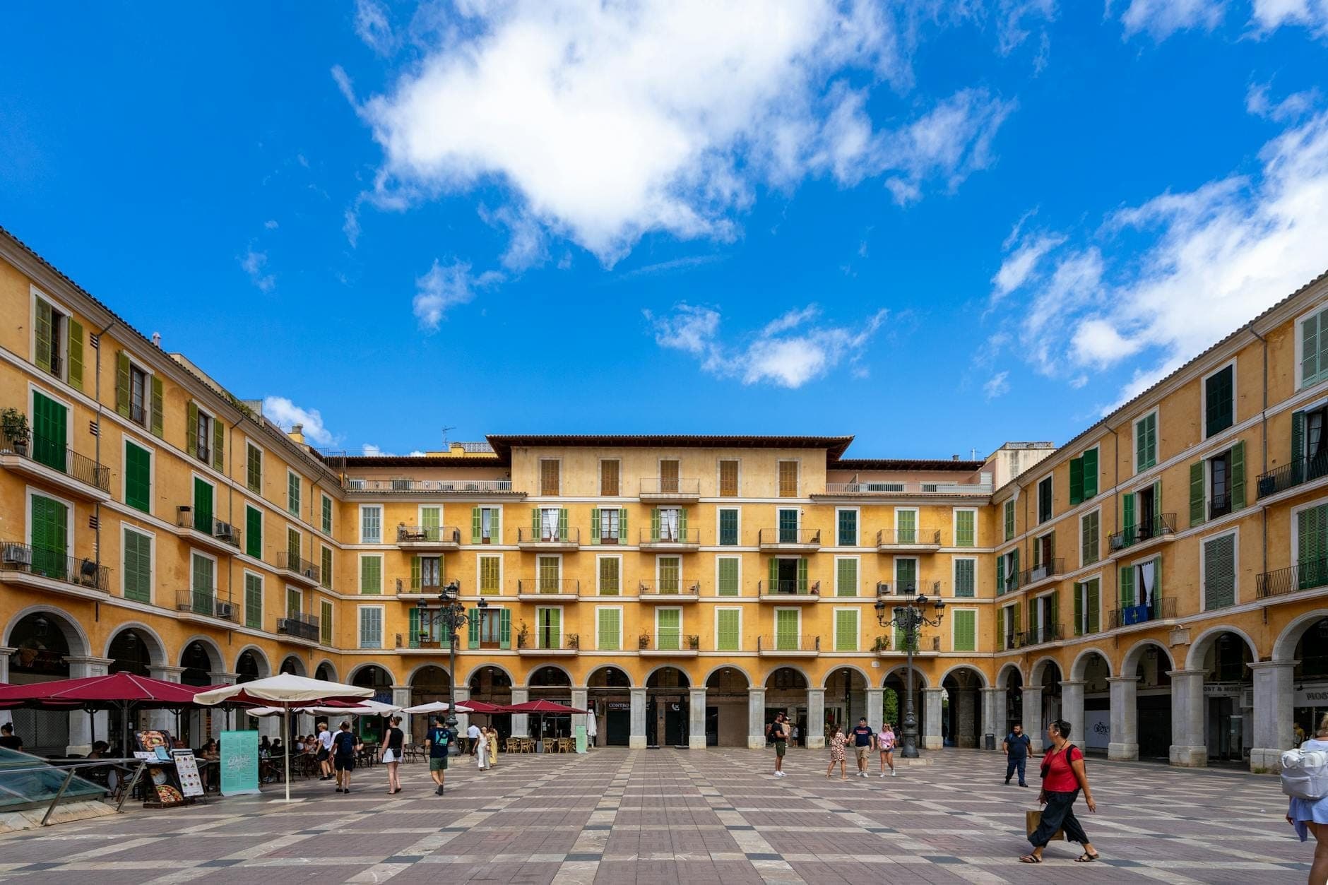 Plaza espaciosa en Palma de Mallorca con edificios amarillos, contraventanas verdes, pasajes con arcos, cafés en la calle y personas paseando bajo un cielo azul.