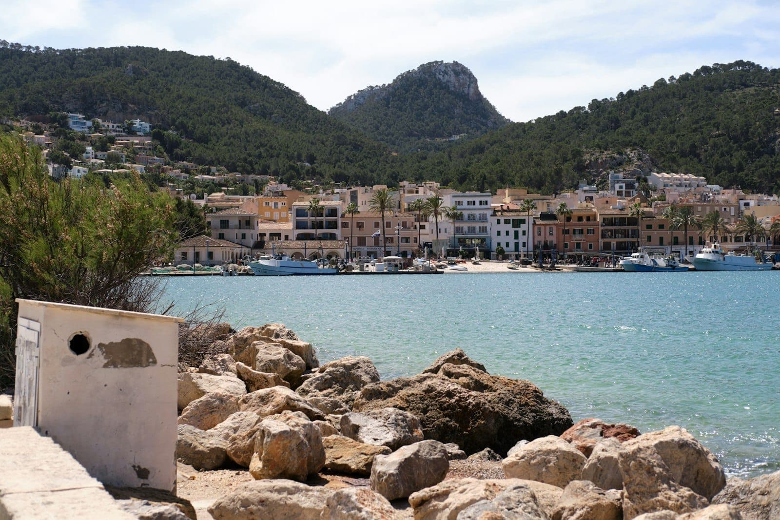 Hafenblick auf Port Andratx auf Mallorca mit bunten Gebäuden, vertäuten Booten, felsiger Küste und grünen Hügeln unter einem strahlenden Himmel.