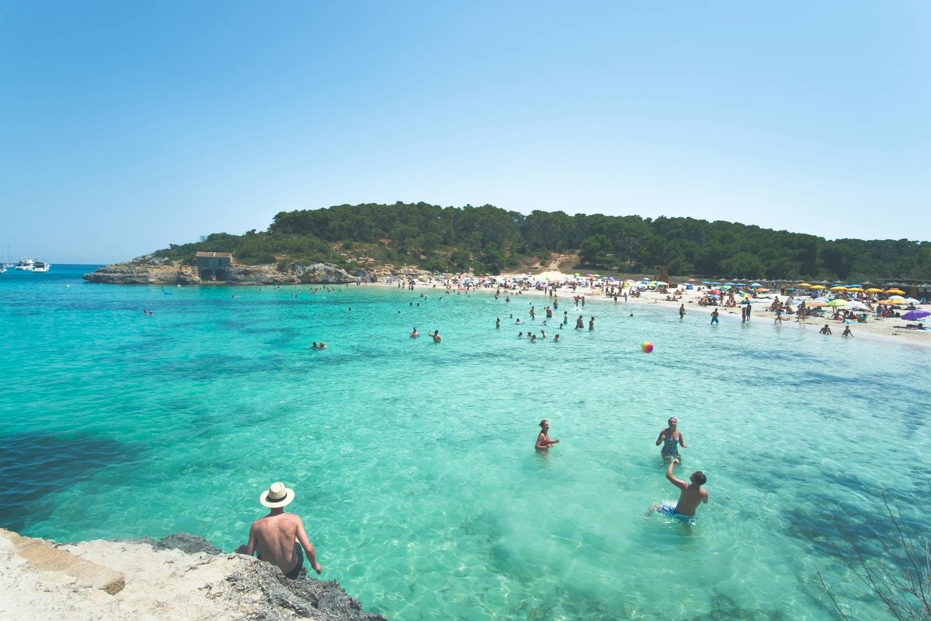 Belebter Strand auf Mallorca im Sommer mit klarem türkisfarbenem Wasser, vielen Menschen beim Schwimmen, Sonnenbaden und Entspannen unter einem sonnigen blauen Himmel.